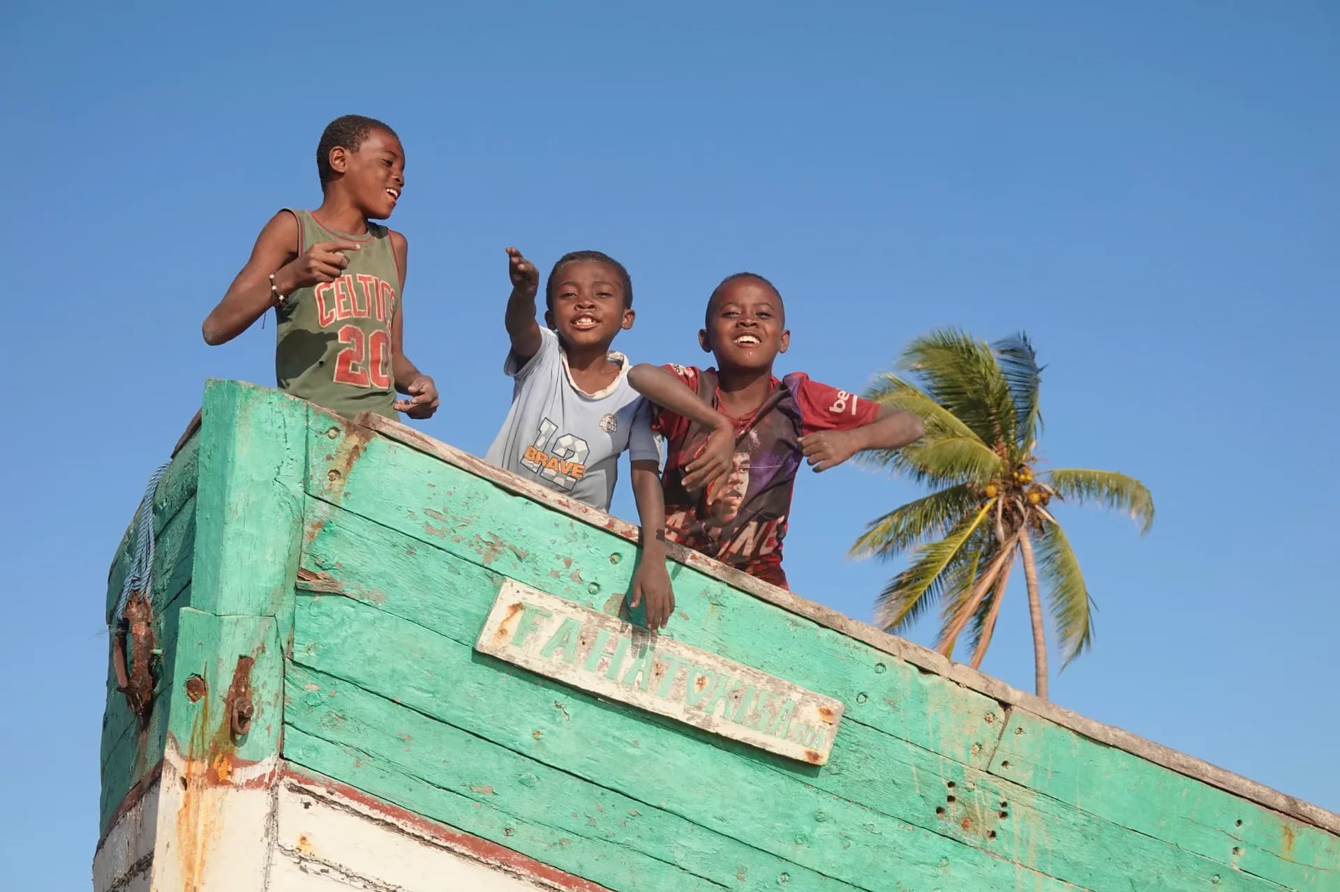 Trois enfants souriants se tiennent dans un bateau coloré, sous un palmier