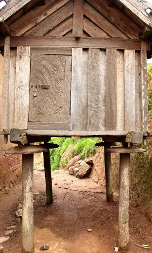 Ancienne cabane en bois sur pilotis avec porte sculptée et sentier naturel en terre