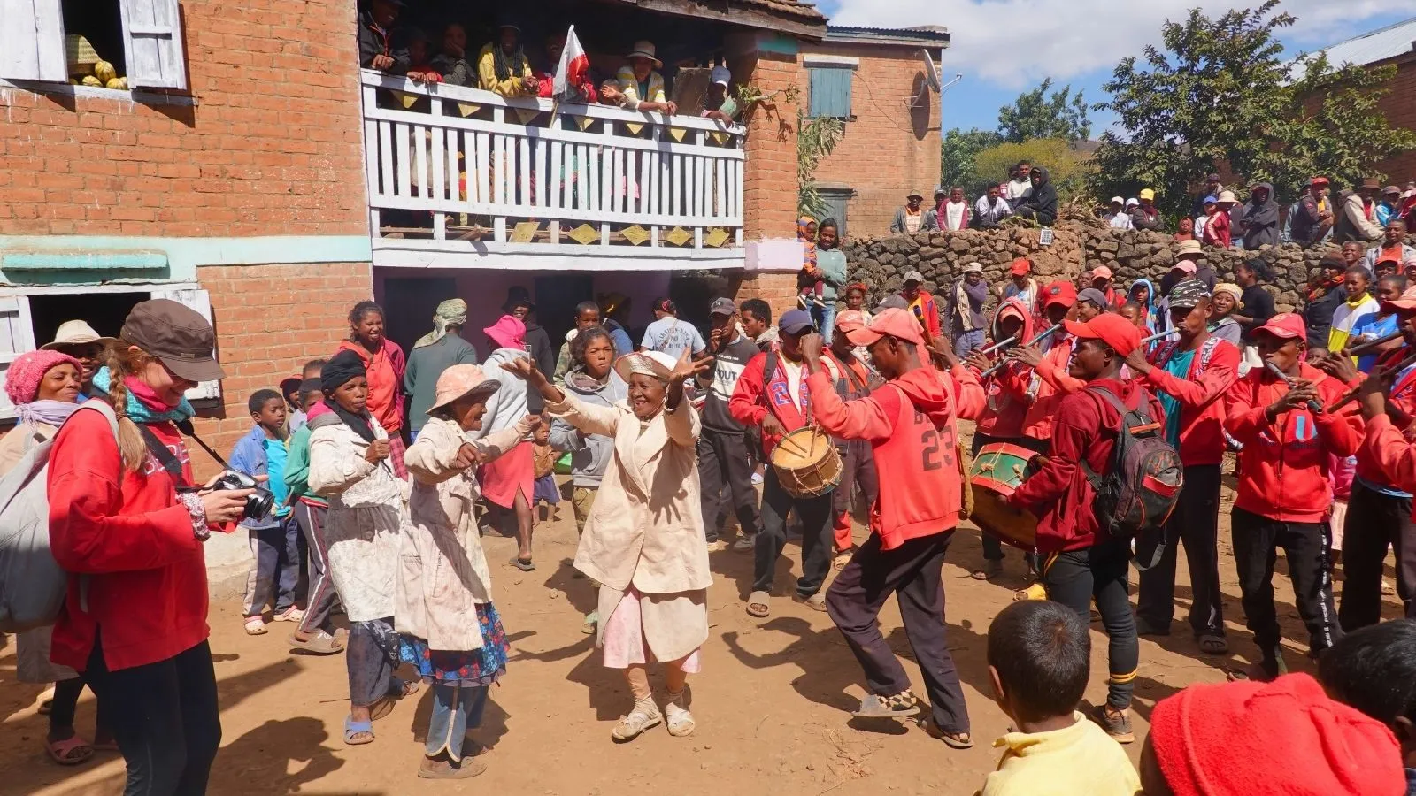 Fête traditionnelle malgache avec danseurs en costumes colorés et spectateurs enthousiastes devant une maison en brique