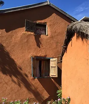 Maison en terre ocre aux volets en bois avec toiture de chaume et fleurs colorées devant