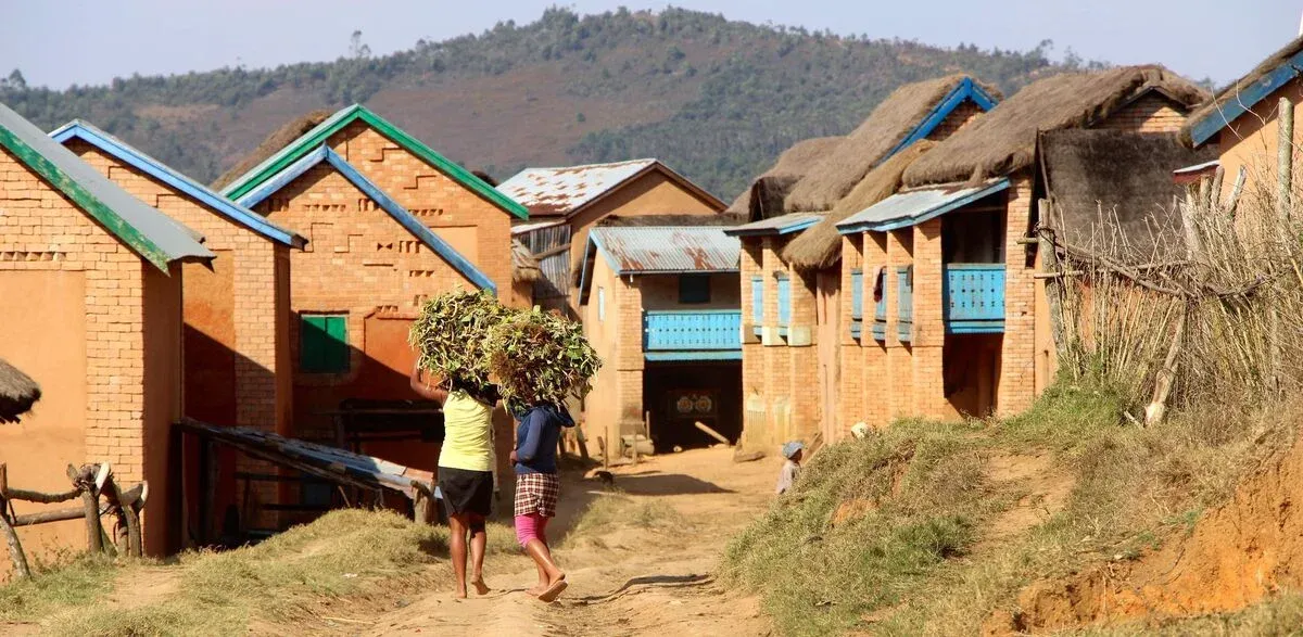Femmes transportant des branches vertes dans un village rural malgache aux maisons colorées et toits de chaume