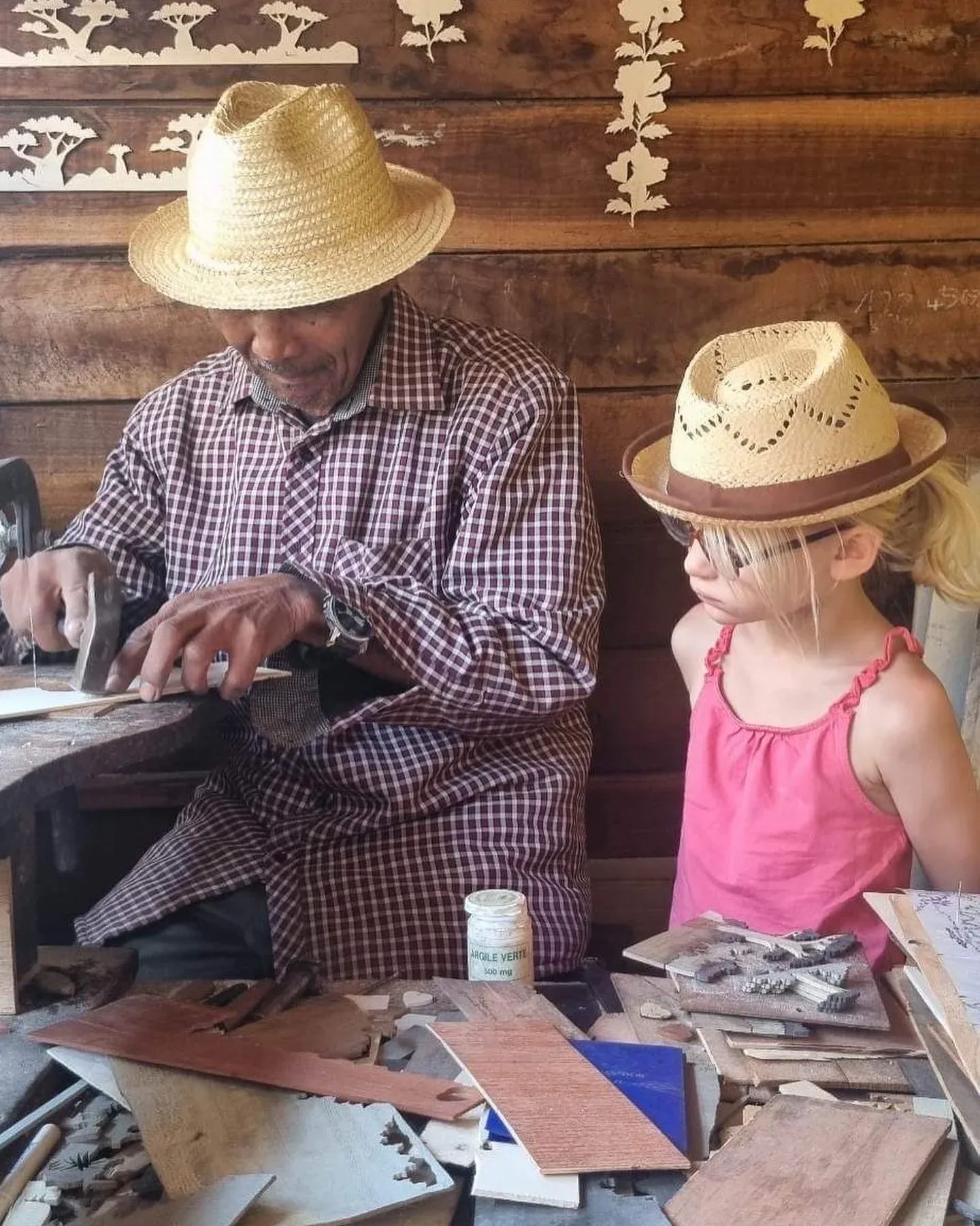 Artisan malgache sculptant des motifs traditionnels devant une jeune spectatrice en chapeau de paille