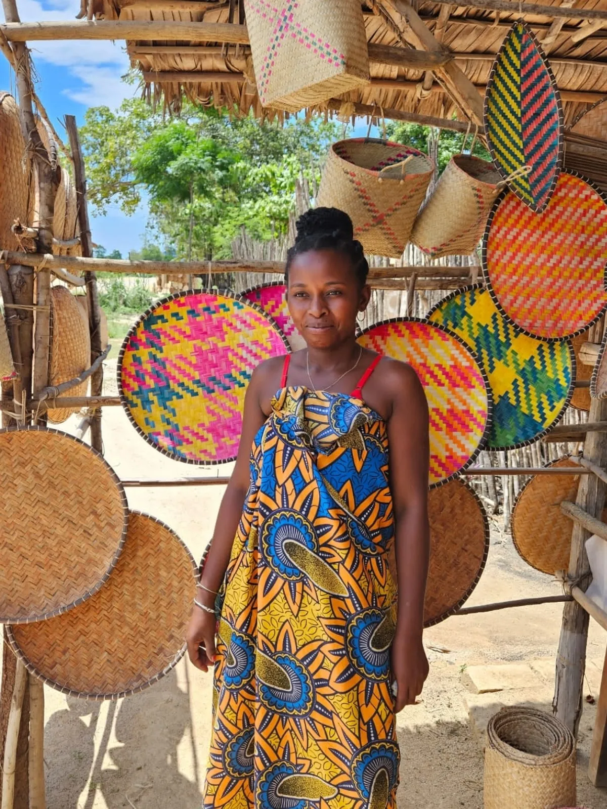 Jeune femme en robe wax colorée devant des paniers et tissages artisanaux malgaches sous abri en paille