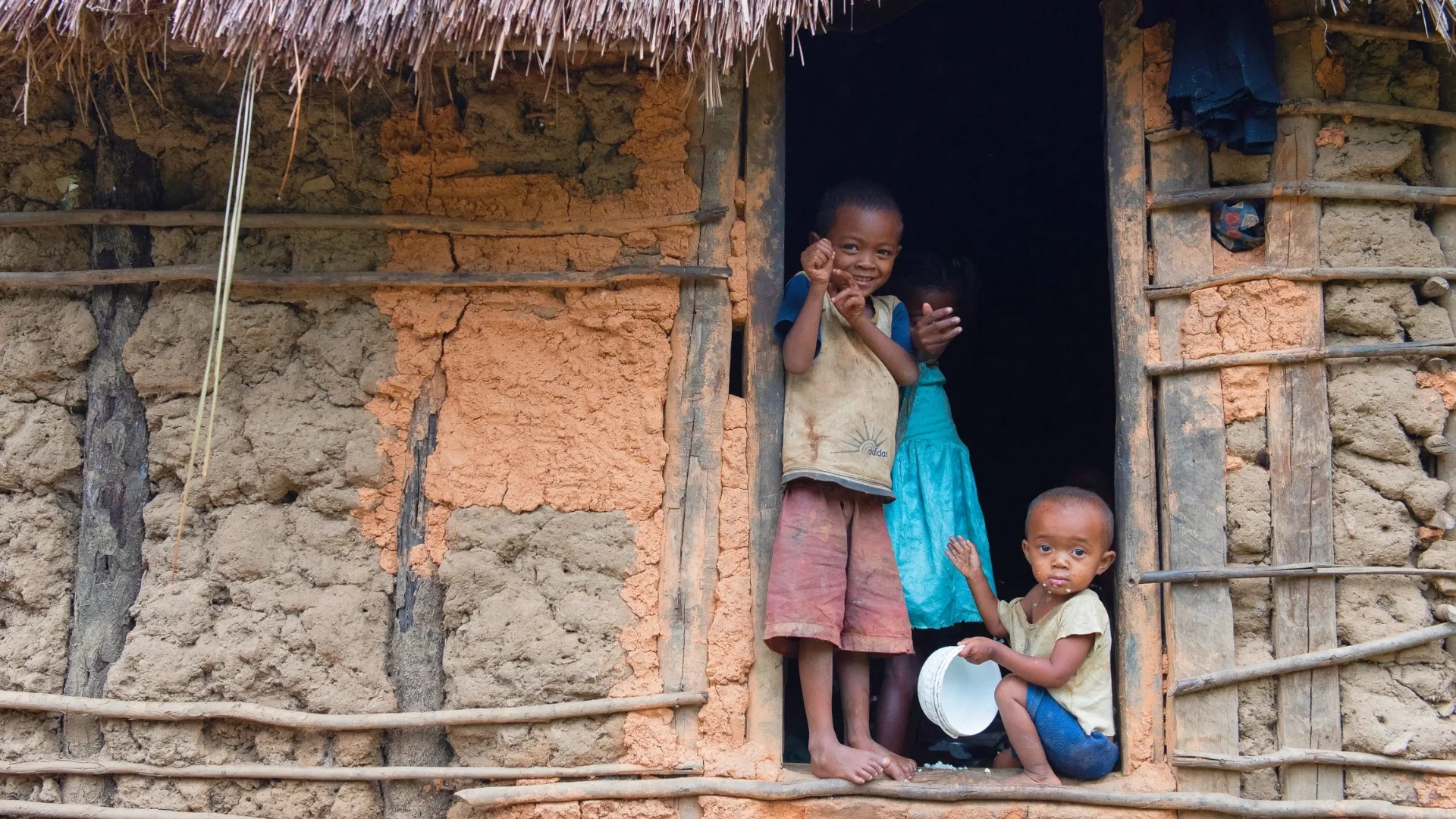 Trois enfants souriants observant depuis l’entrée d’une modeste maison en terre de Madagascar