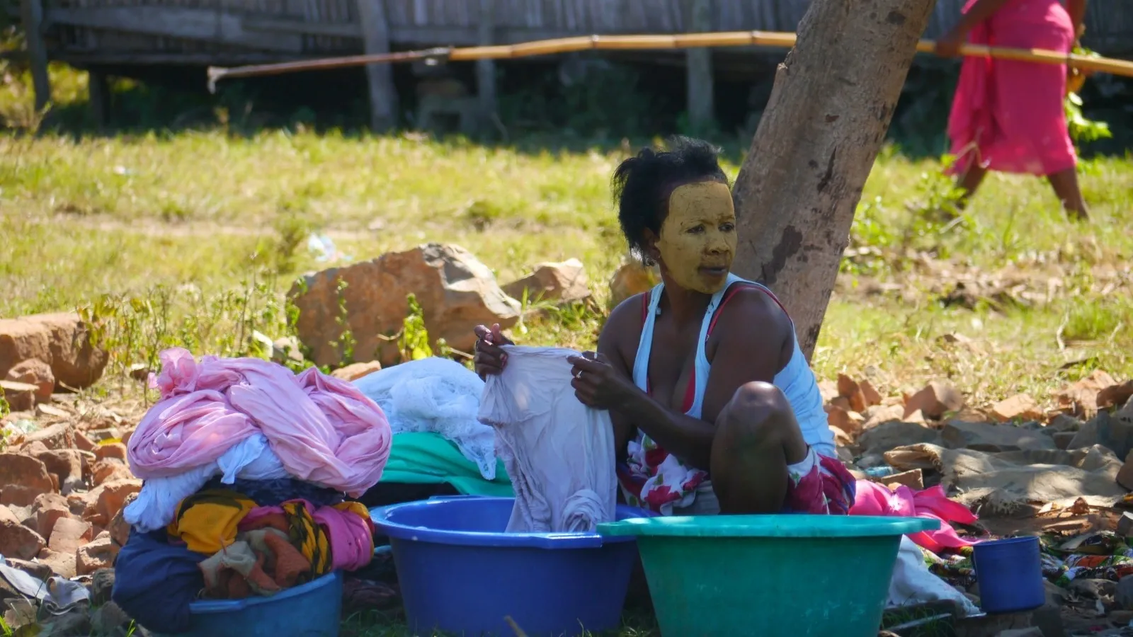 Femme assise près d’un arbre lavant des vêtements colorés dans des bassines en plastique