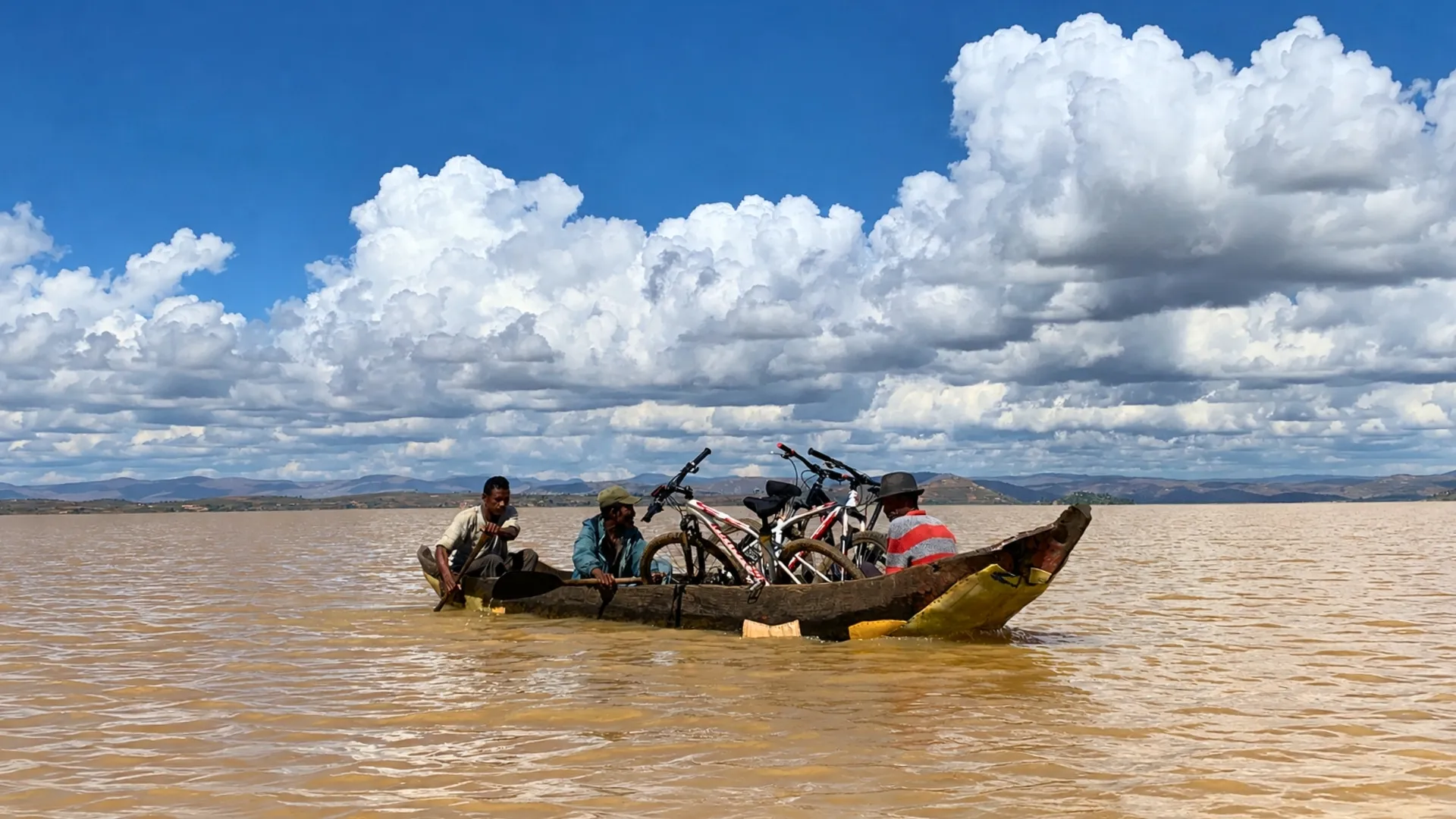 Trois personnes transportant des vtt dans une pirogue sur un fleuve malgache sous un ciel nuageux