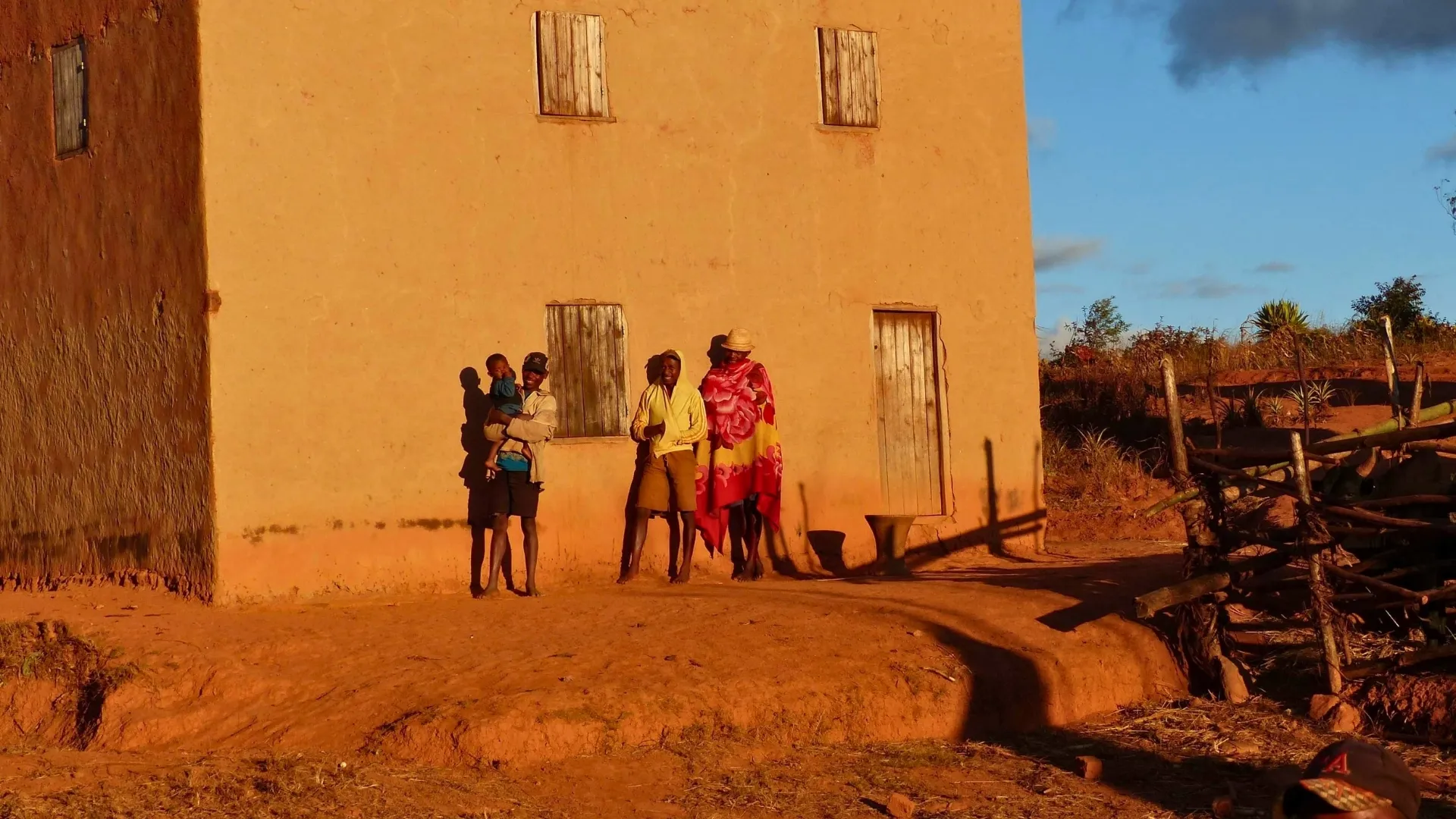 Groupe familial devant une maison en terre sous lumière dorée en région rurale malgache