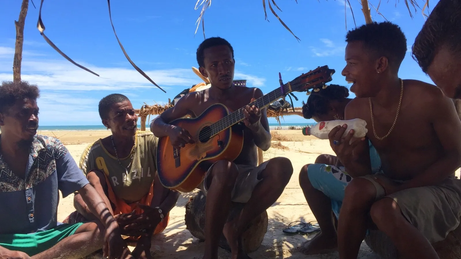 Groupe détendu sur plage sous abri naturel jouant de la guitare et discutant joyeusement