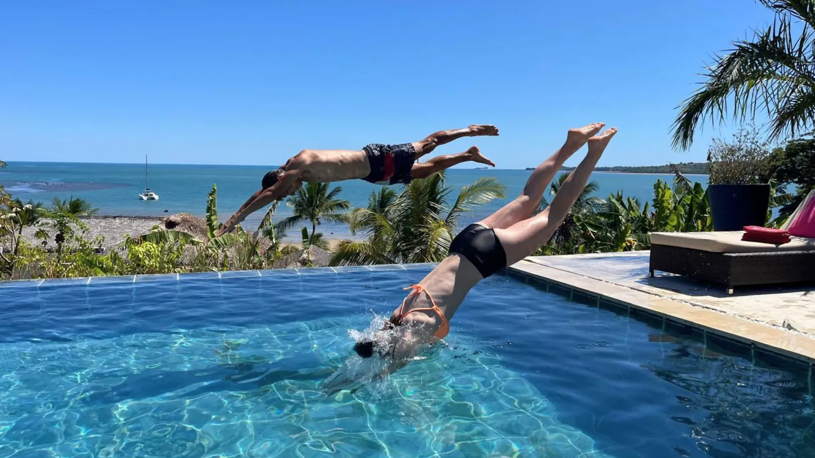 Deux plongeurs sautant dans une piscine à débordement face à une mer turquoise et des palmiers